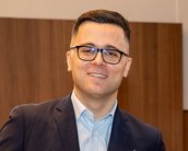 Ein Mann mit Brille und Anzug lächelt in einem modernen Raum. Er hat kurze Haare und steht leicht zur Seite.