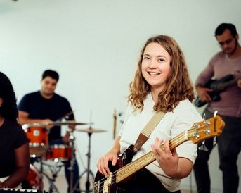 Eine Frau mit einer E-Gitarre lächelt in die Kamera, während andere Musiker im Hintergrund spielen.