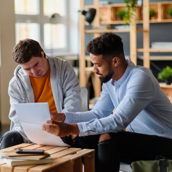 Zwei Männer sitzen an einem Tisch und besprechen Dokumente. Einer hält Papiere, während der andere aufmerksam zuhört.