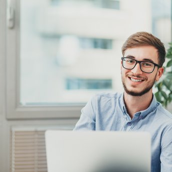 A young man with glasses is sitting at a desk and smiling. He is wearing a light blue shirt and working on a laptop. In the background, there are windows and a plant visible.