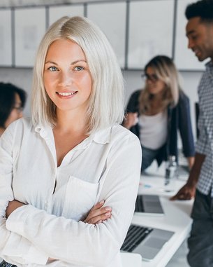 Eine blonde Frau mit schulterlangen Haaren steht in einem modernen Büro und lächelt, während sie die Arme verschränkt. Im Hintergrund sind drei weitere Personen zu sehen, die an Tischen arbeiten und miteinander sprechen.