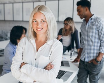 Eine blonde Frau mit schulterlangen Haaren steht in einem modernen Büro und lächelt, während sie die Arme verschränkt. Im Hintergrund sind drei weitere Personen zu sehen, die an Tischen arbeiten und miteinander sprechen.
