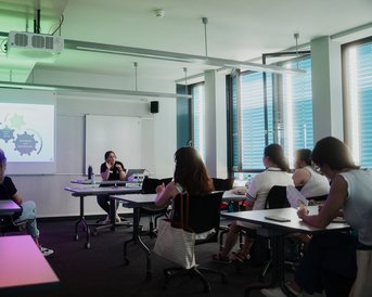 A person is presenting in front of a group of listeners in a modern classroom, while a slide with diagrams is displayed in the background.