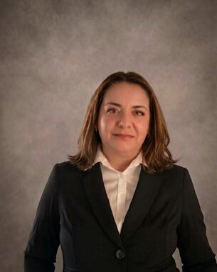 Eine Frau mit langen, braunen Haaren trägt einen schwarzen Blazer und ein weißes Hemd. Der Hintergrund ist grau und unscharf.