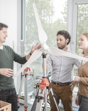 Drei Personen stehen in einem Raum und betrachten ein Modell einer Windturbine. Eine Person erklärt die Funktionsweise, während die anderen aufmerksam zuhören. Werkzeuge und Materialien sind im Hintergrund sichtbar.