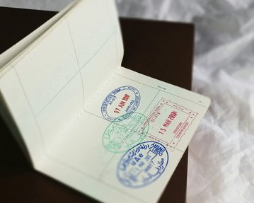 A opened passport book with several stamps, including a red stamp and two blue stamps representing various entry or exit stamps. The background is blurred and neutral.