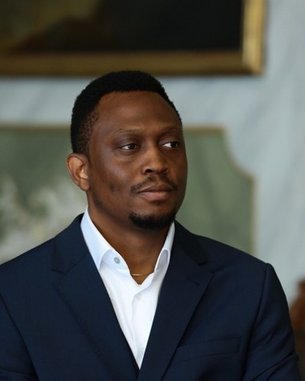 A man in a suit sits with a neutral expression. He has short, curly hair and is wearing a white shirt under a dark blazer.