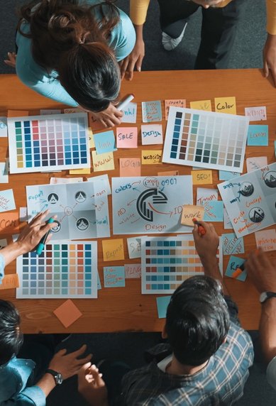 Several people are working at a table, surrounded by color samples, notes, and a laptop, as they exchange ideas.