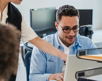 Eine Person zeigt einem Mann am Computer etwas, während er aufmerksam schaut. Beide sind in einem modernen Büro.