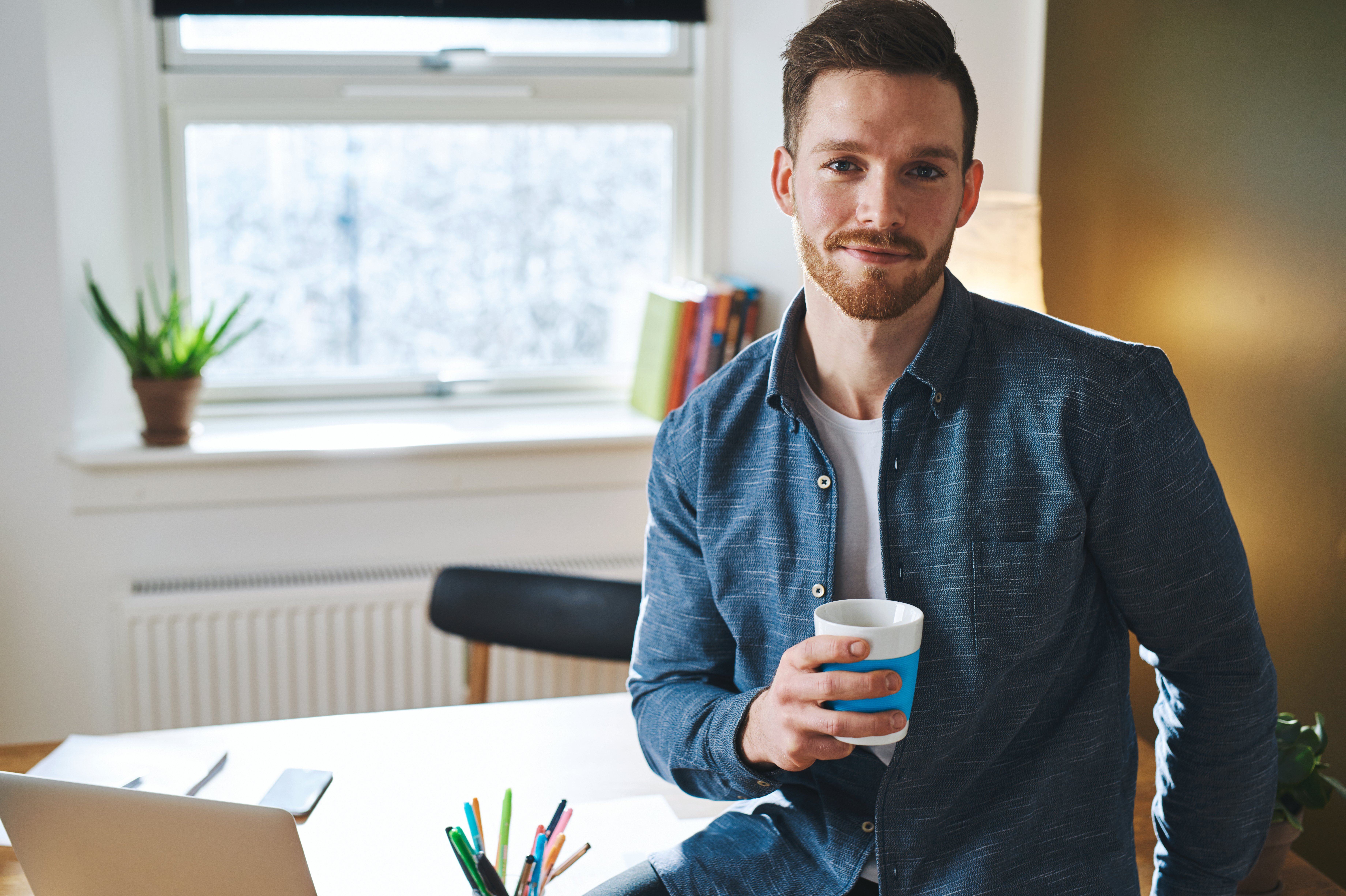 Ein Mann mit einem Bart hält einen Kaffeebecher in der Hand. Er trägt ein blaues Hemd und sitzt an einem Schreibtisch mit einem Laptop und Schreibutensilien. Im Hintergrund sind Pflanzen und Bücherregale sichtbar.