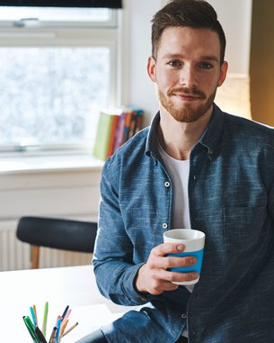 Ein Mann mit einem Bart hält einen Kaffeebecher in der Hand. Er trägt ein blaues Hemd und sitzt an einem Schreibtisch mit einem Laptop und Schreibutensilien. Im Hintergrund sind Pflanzen und Bücherregale sichtbar.