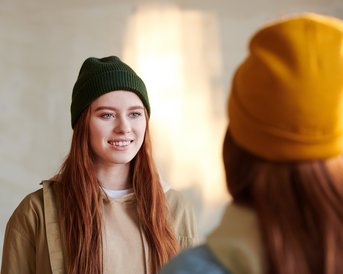 Eine junge Frau mit langen, roten Haaren trägt eine grüne Mütze und lächelt. Sie steht in einem hellen Raum und spricht mit einer anderen Person, die eine gelbe Mütze trägt.