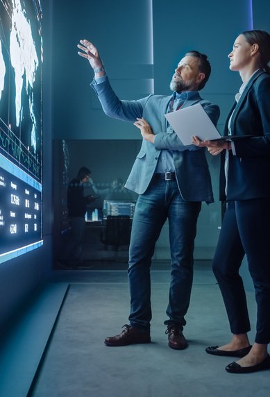 A man and a woman are standing in front of a large digital display featuring graphics and data analyses, while they engage in a discussion.