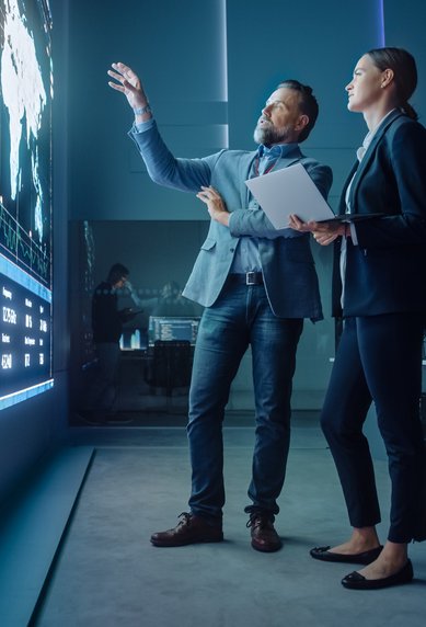 Ein Mann und eine Frau stehen vor einer großen digitalen Anzeige mit Grafiken und Datenanalysen, während sie eine Diskussion führen.