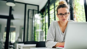 A woman with glasses is sitting at a table, focused on working on a laptop in a bright, modern room.