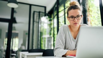 Eine Frau mit Brille sitzt an einem Tisch und arbeitet konzentriert an einem Laptop in einem hellen, modernen Raum.