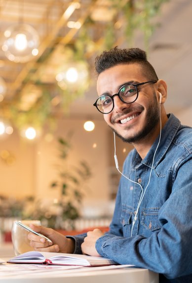 A young man with glasses is sitting at a table in a café, working on his laptop while holding a notebook and a smartphone.