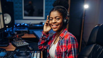 Eine junge Frau mit Kopfhörern lächelt am DJ-Pult, umgeben von Musikgeräten und einem Computerbildschirm.
