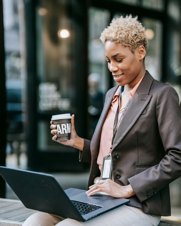 A woman with short, curly hair is sitting at a table, working on a laptop. She is holding a coffee mug in one hand and is wearing a blazer along with an ID badge around her neck.