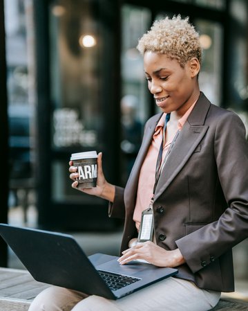 Eine Frau mit kurzen, lockigen Haaren sitzt an einem Tisch und arbeitet an einem Laptop. Sie hält einen Kaffeebecher in der Hand und trägt einen Blazer sowie eine ID-Karte um den Hals.