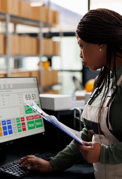 A woman in a work suit stands at a computer, checking data on the screen while holding documents in her hand.