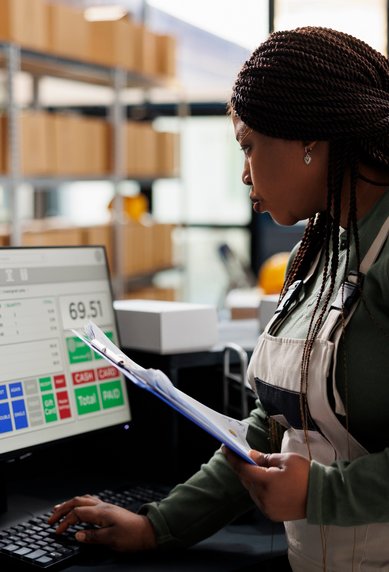 Eine Frau in einem Arbeitsanzug steht an einem Computer und überprüft Daten auf dem Bildschirm, während sie Unterlagen in der Hand hält.