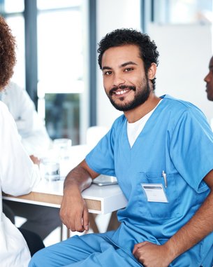 Ein junger Mann in blauer medizinischer Kleidung sitzt an einem Tisch und lächelt. Um ihn herum sind mehrere Personen in ähnlicher Kleidung, die an einer Besprechung teilnehmen. Der Raum hat große Fenster mit viel natürlichem Licht.