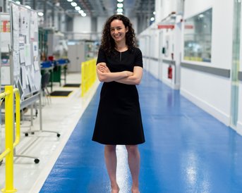 A woman with curly hair stands in a modern, bright production area. She is wearing a black dress and has her arms crossed. The floor is blue, and in the background, machines and workstations are visible.