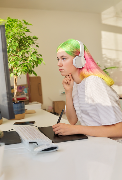 Eine Person mit bunten Haaren sitzt an einem Schreibtisch, trägt Kopfhörer und schaut konzentriert auf einen Computerbildschirm.