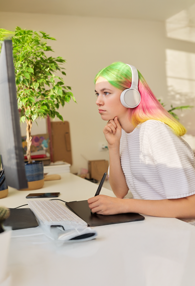 A person with colorful hair is sitting at a desk, wearing headphones and intently looking at a computer screen.