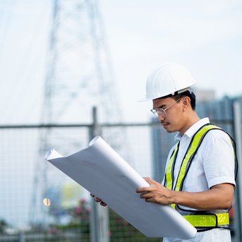 Ein Bauarbeiter in einem Sicherheitshelm und einer Warnweste betrachtet Baupläne vor einem Zaun und einem Hochspannungsmast.