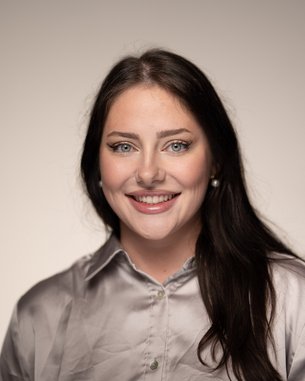 Eine lächelnde Frau mit langen, dunklen Haaren trägt ein graues Hemd. Der Hintergrund ist neutral und hell.