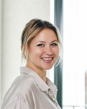 Eine lächelnde Frau mit blonden Haaren trägt ein helles Hemd und steht vor einem Fenster mit natürlichem Licht.