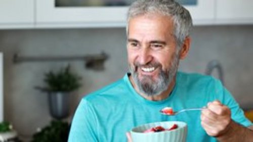Ein lächelnder älterer Mann mit grauem Bart hält eine Schüssel mit Obst und einen Löffel in der Hand, während er in einer modernen Küche sitzt.