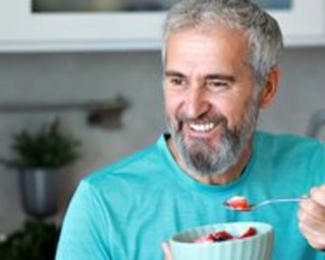 A smiling older man with a gray beard holds a bowl of fruit and a spoon in his hand while sitting in a modern kitchen.