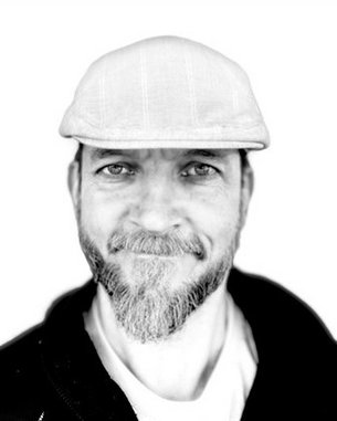 Ein Mann mit einem Bart und einer Schirmmütze lächelt in die Kamera. Der Hintergrund ist unscharf und hell.