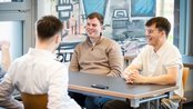 Drei junge Männer sitzen an einem Tisch und unterhalten sich. Einer trägt einen beigen Pullover, die anderen Hemden.