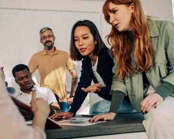 Eine Gruppe von fünf Personen diskutiert und arbeitet an einem Tisch, während sie Dokumente und Notizen betrachten.
