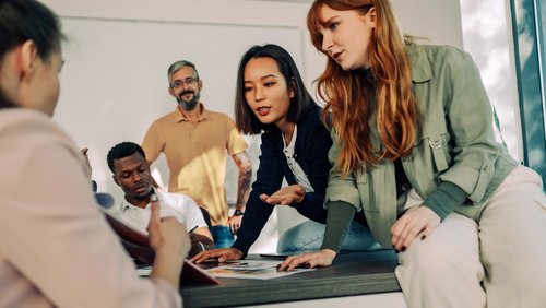 Eine Gruppe von fünf Personen diskutiert und arbeitet an einem Tisch, während sie Dokumente und Notizen betrachten.