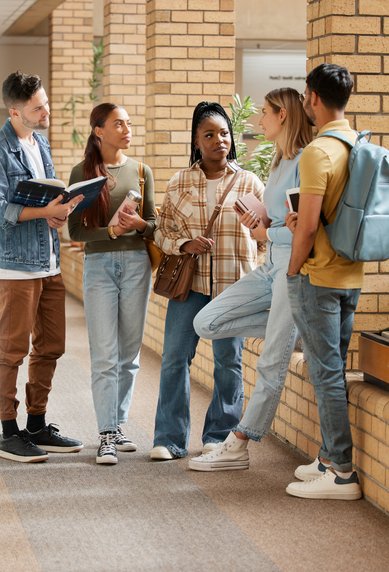 Five students are standing in a hallway, discussing. They are holding books and notes as they talk.