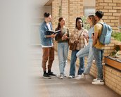 Five students are standing in a hallway, discussing. They are holding books and notes as they talk.