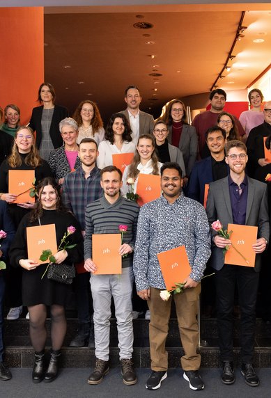 Gruppenszene mit mehreren Personen, die auf einer Treppe stehen. Die Teilnehmer halten orangefarbene Urkunden und Blumen in den Händen. Die Gruppe besteht aus Männern und Frauen unterschiedlichen Alters.
