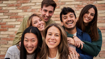 Six people are smiling together in a group in front of a brick wall. They are standing close to each other, creating a friendly and relaxed atmosphere.