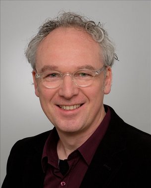 Ein Mann mit lockigem, grauem Haar und Brille lächelt freundlich. Er trägt ein schwarzes Sakko über einem burgunderfarbenen Hemd. Der Hintergrund ist neutral und hell.
