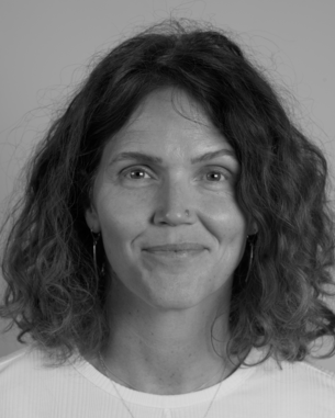 Eine Person mit lockigem, schulterlangem Haar und Ohrhängern lächelt in die Kamera vor einem neutralen Hintergrund.