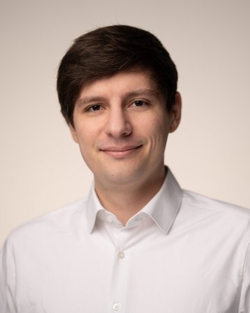 A young man with brown hair is wearing a white shirt and smiling warmly at the camera against a neutral background.