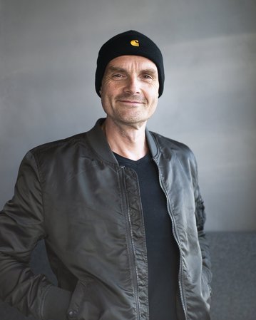 Mann mit lässiger Jacke und schwarzer Mütze steht lächelnd vor einem neutralen Hintergrund.