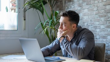 A man sits at a table, thoughtfully looking at a laptop with his hand on his chin. In the background, a plant is visible.