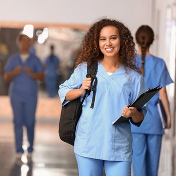 Eine junge Frau in blauer Arbeitskleidung hält eine Mappe und lächelt, während sie in einem Krankenhausflur steht. Im Hintergrund sind weitere Personen in ähnlicher Kleidung zu sehen.