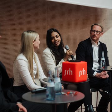 Drei Personen sitzen an einem Tisch, während eine Frau mit einem Mikrofon spricht. Auf dem Tisch steht ein roter Würfel mit der Aufschrift "ssrh".